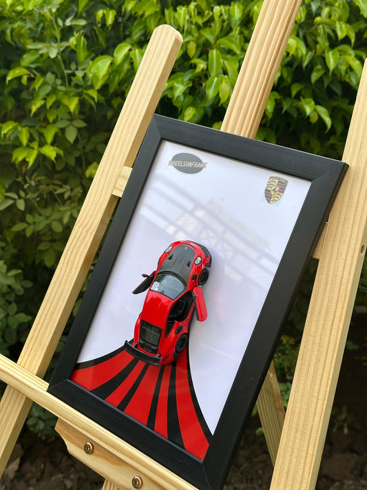 Framed artwork of a red porsche 911 car on a wooden easel with green foliage in the background