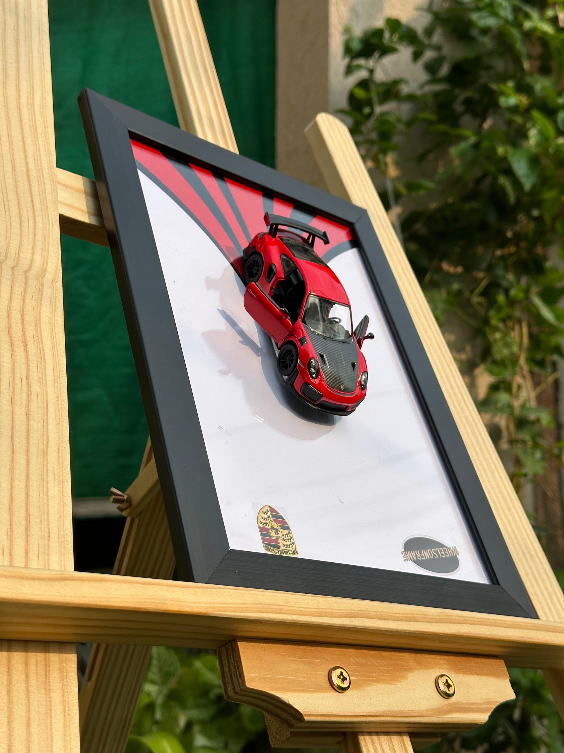Framed picture of a red porsche 911 diecast model sports car on a wooden easel with a blurred green background