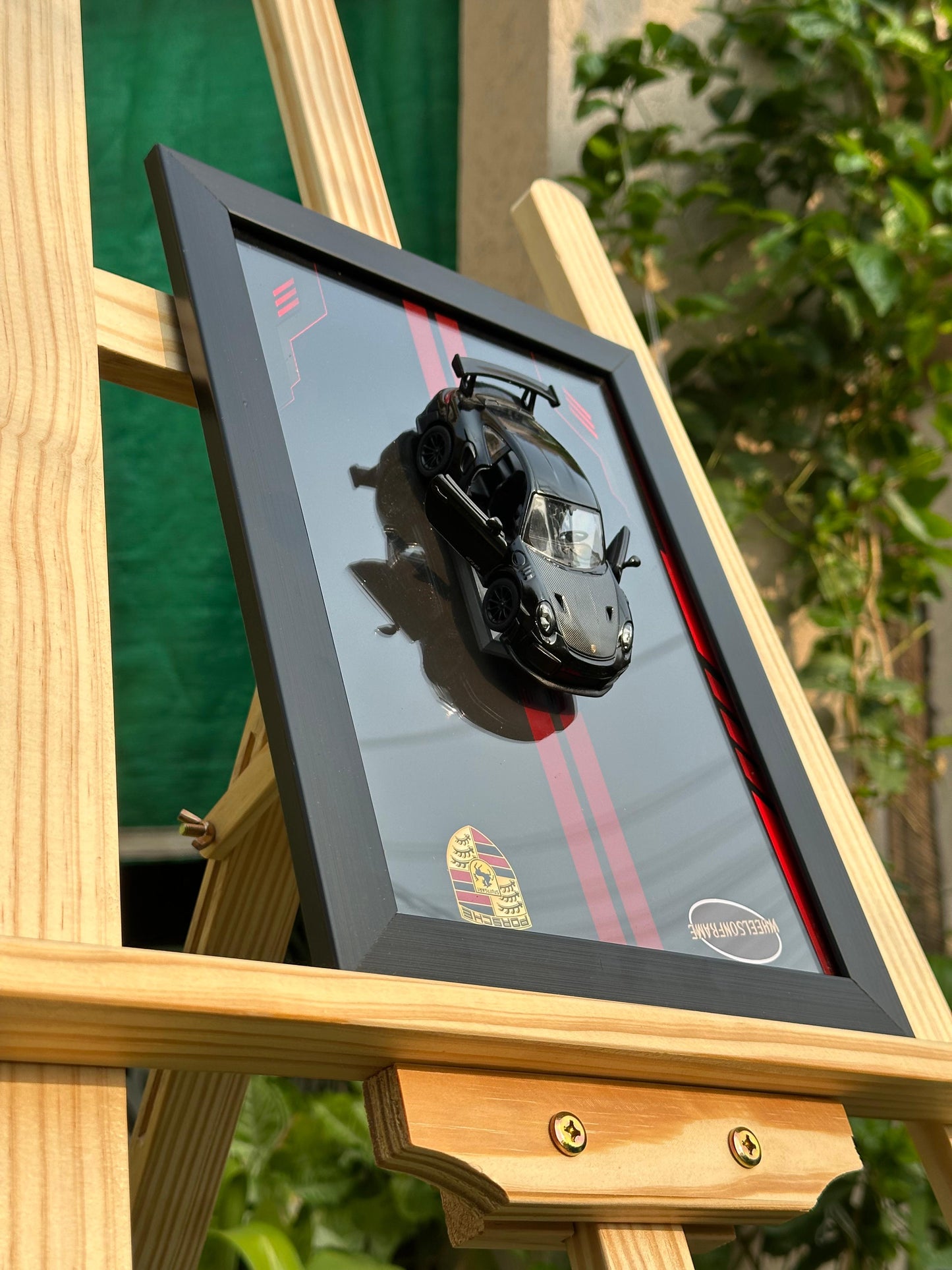 Framed artwork of a porsche 911 diecast car model on a wooden easel with a blurred green background