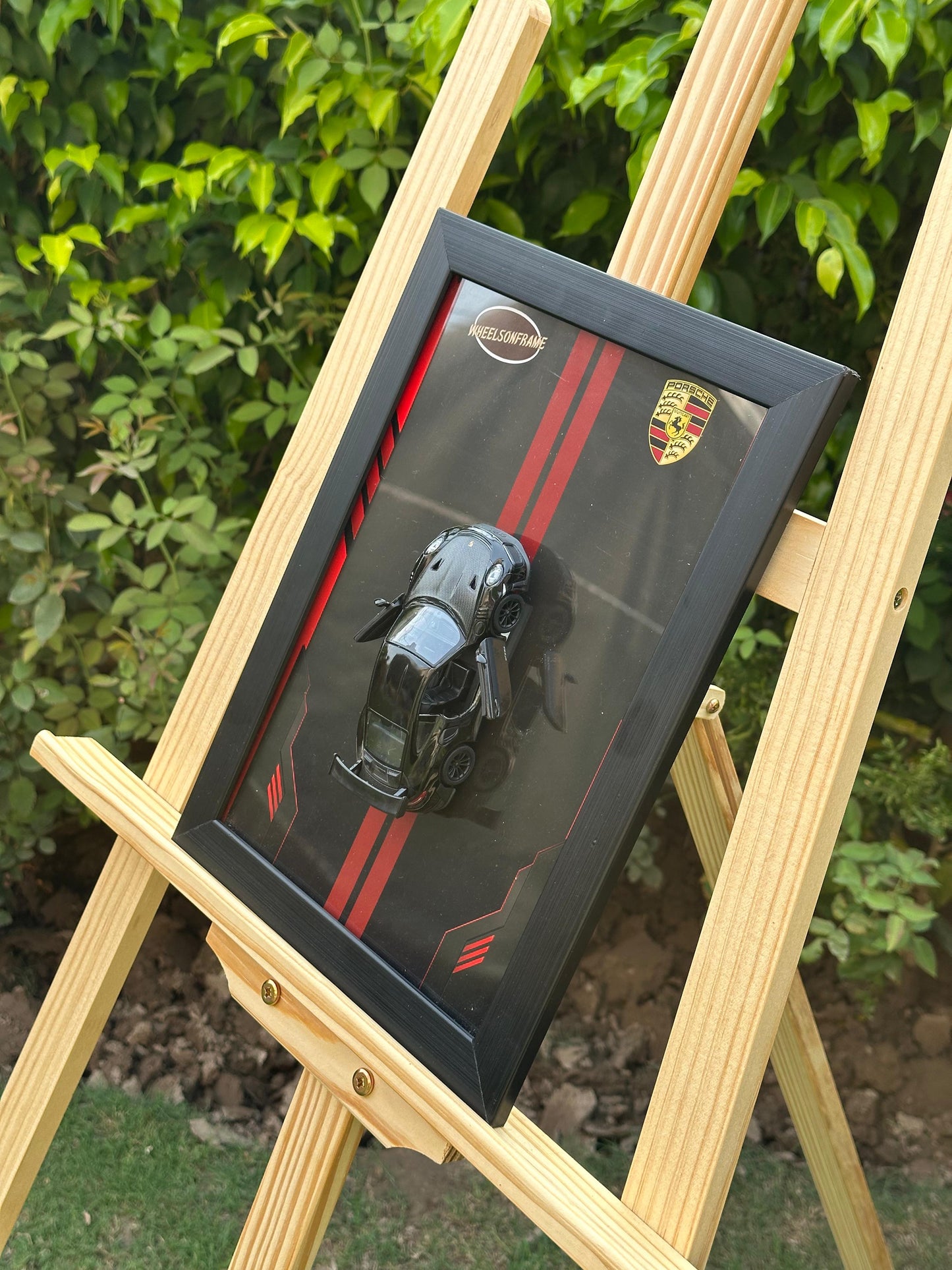 Framed picture of a black porsche 911 on a wooden easel with a blurred natural background