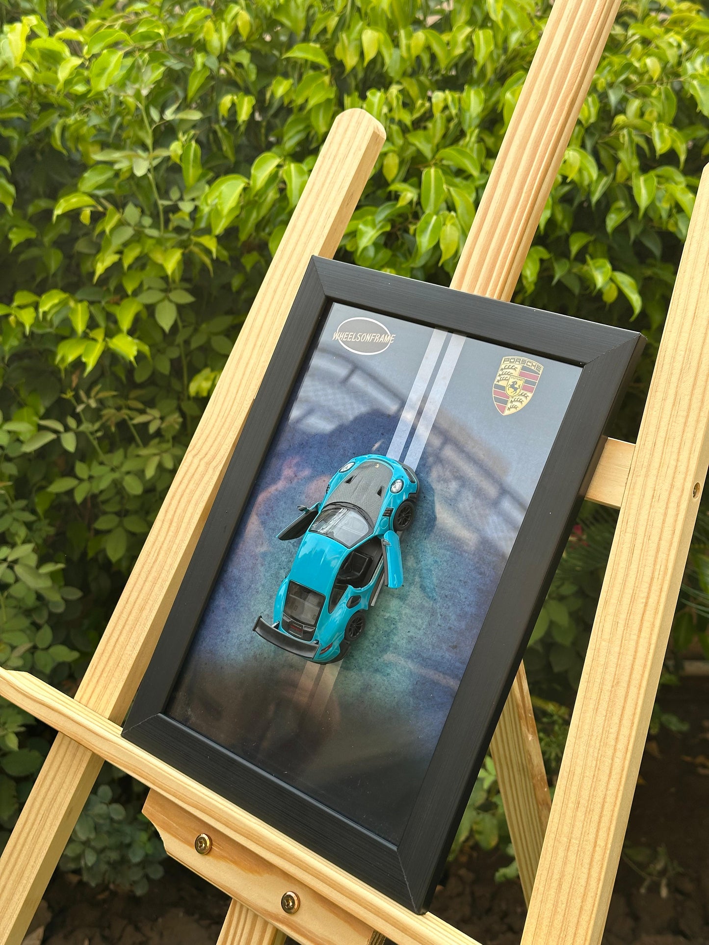 Framed model picture of a blue Porsche 911 sports car on a wooden easel with green foliage in the background